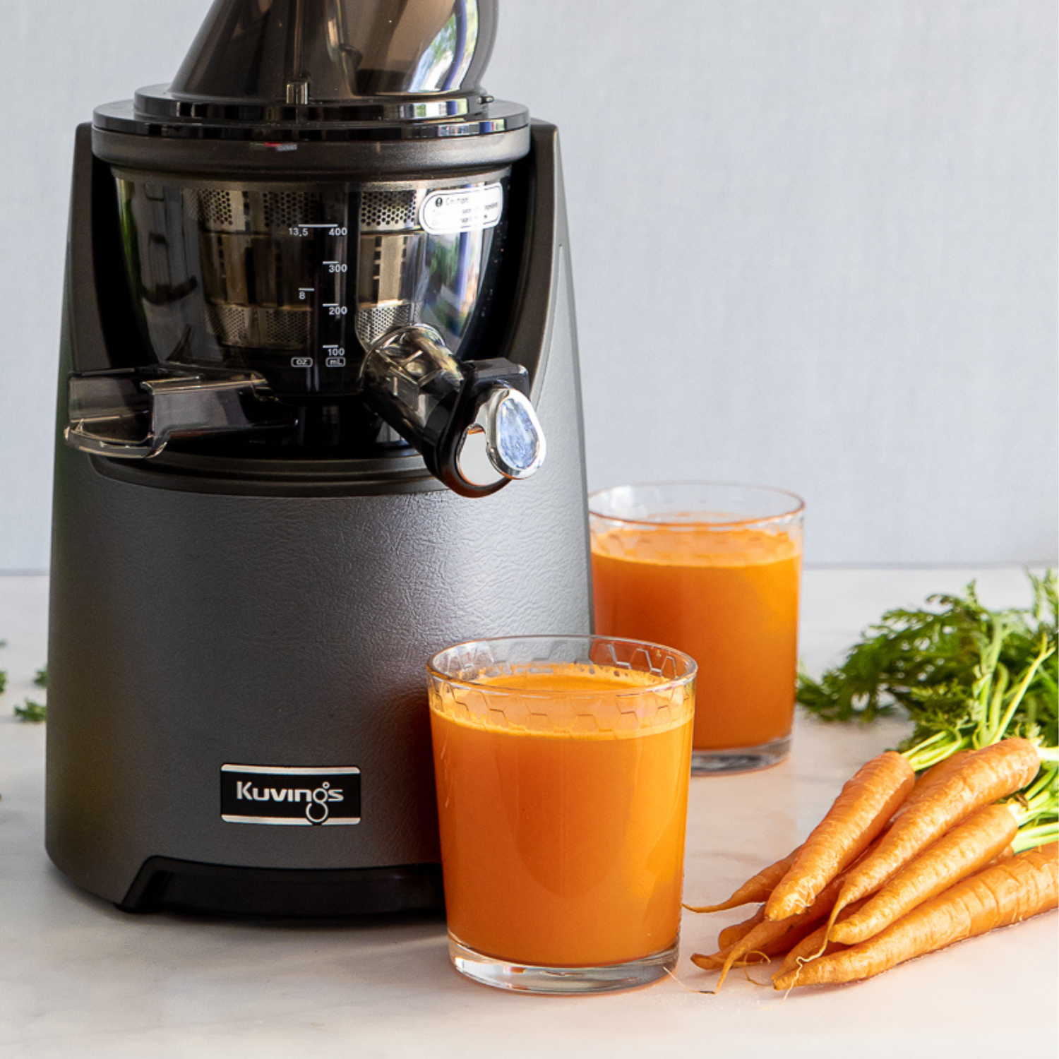 Kuvings EVO820 juicer next to 2 glasses of orange-colored juice and a bunch of carrots.