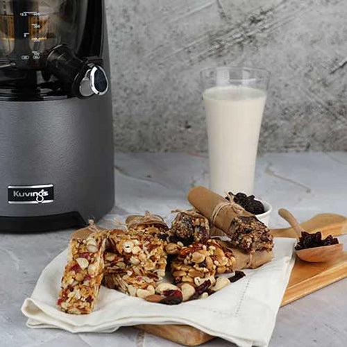 Nut pulp energy bars stacked on a wooden board in front of a glass of milk and a Kuvings juicer.