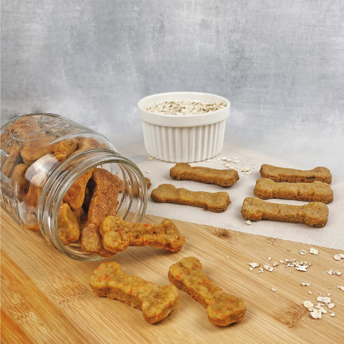 Bone-shaped dog treats spilling out of a tipped jar, next to a ramekin of oats. 