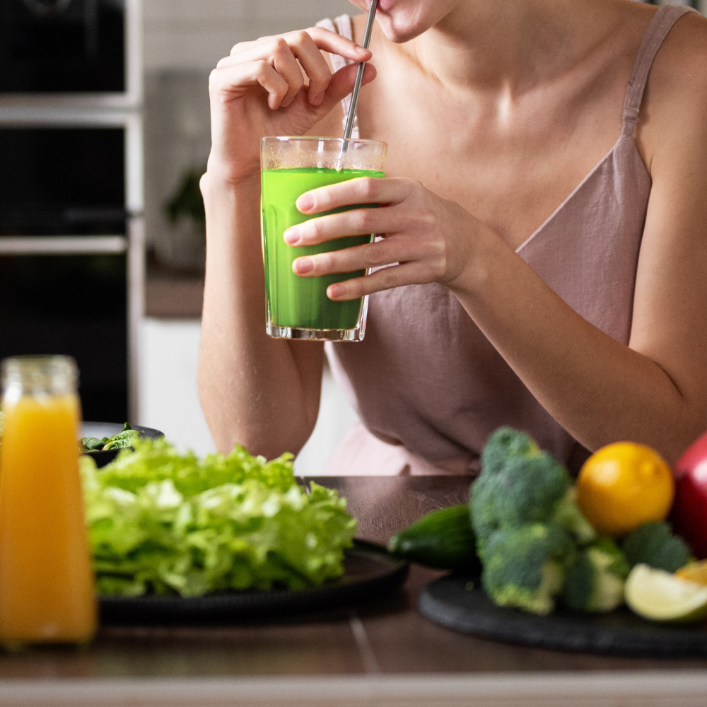 Woman drinking a glass of green juice over a table of fresh fruits and vegetables