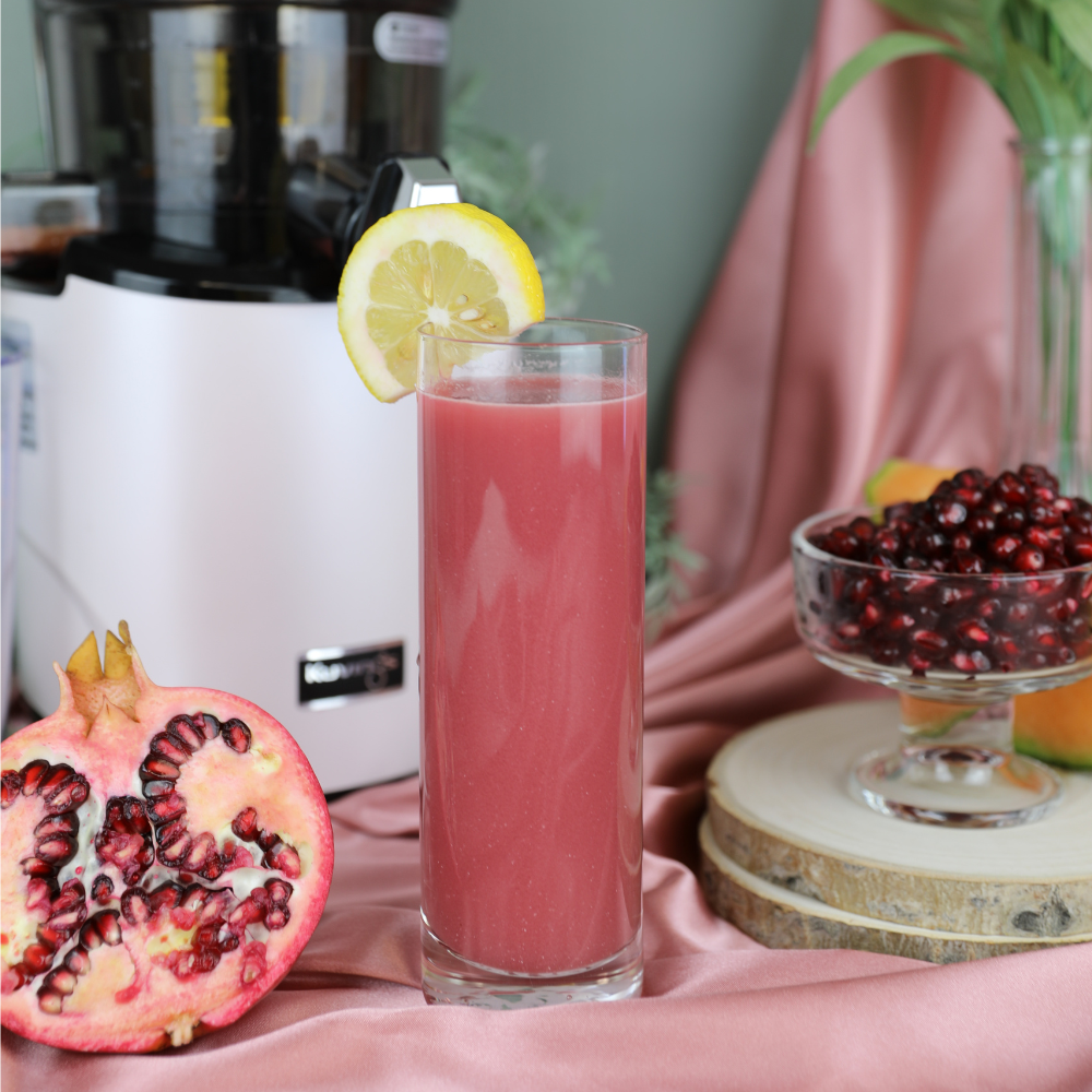 Pink juice in a tall glass with a lemon slice. Pomegranate and cantaloupe in the background with a Kuvings juicer.