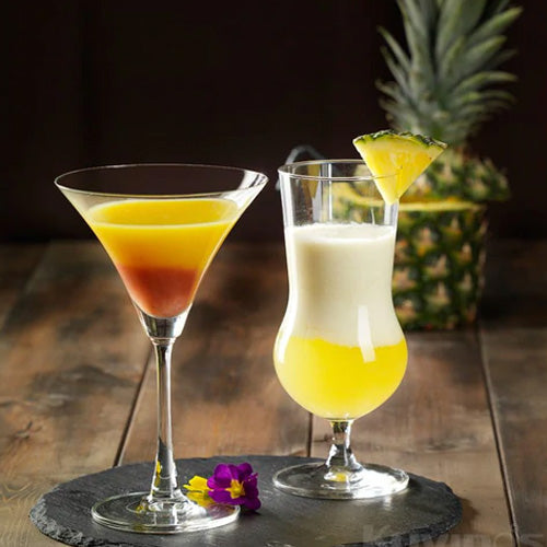 2 cocktails on wood table in front of whole pineapple