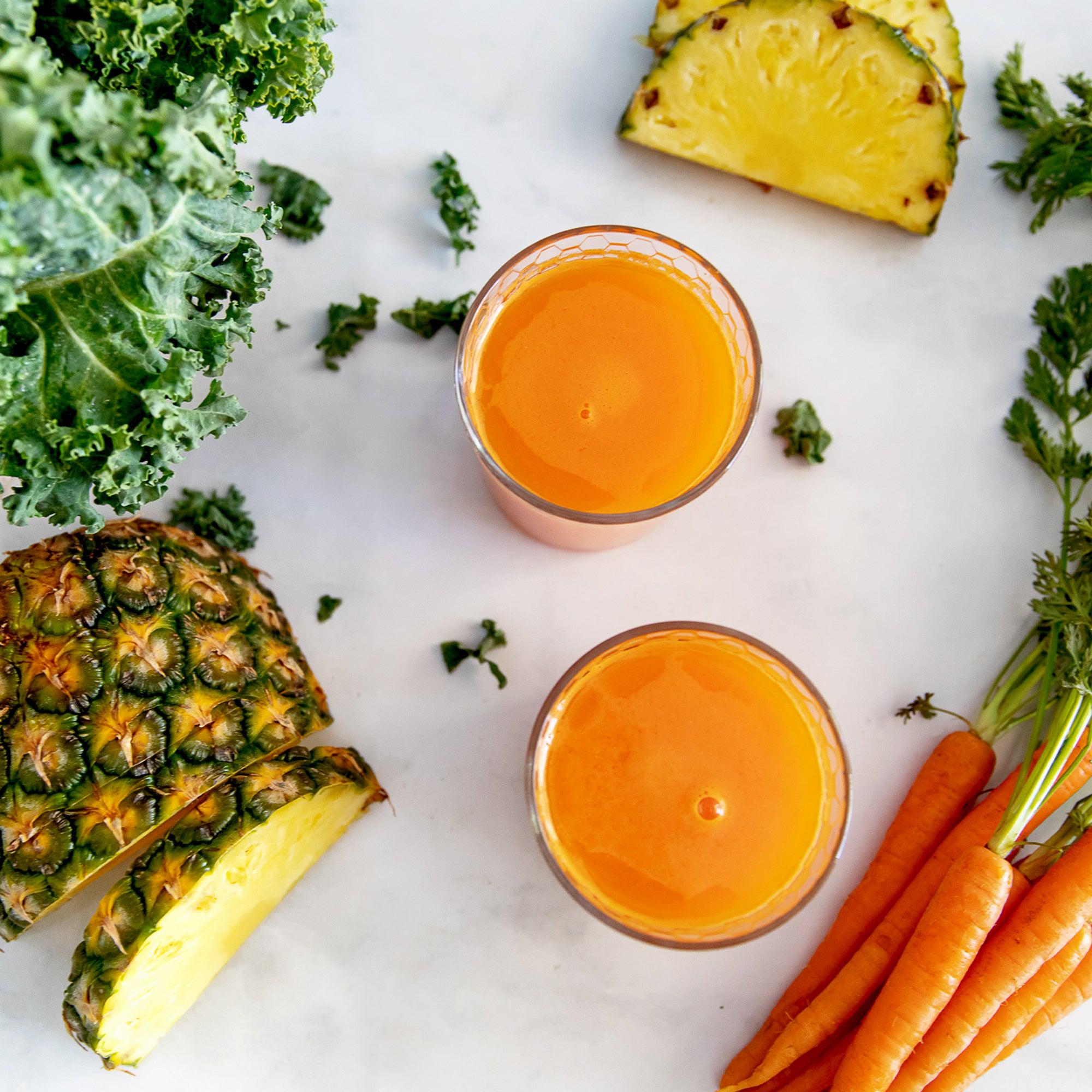 Two glasses of carrot juice surrounded by carrots, pineapple slices, and kale.