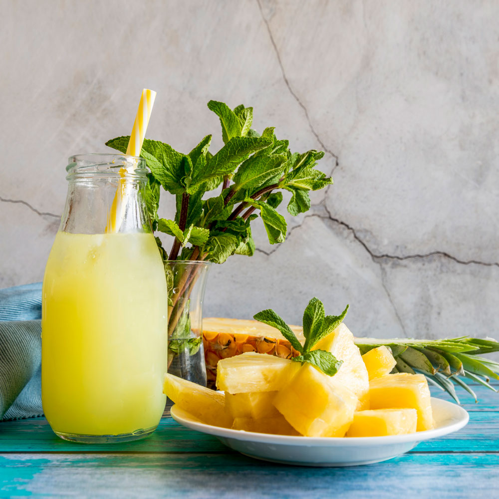 spring pineapple juice in a glass with a straw. slices of pineapple on a plate. half a pineapple. mint leaves in a vase with water on a blue wood table